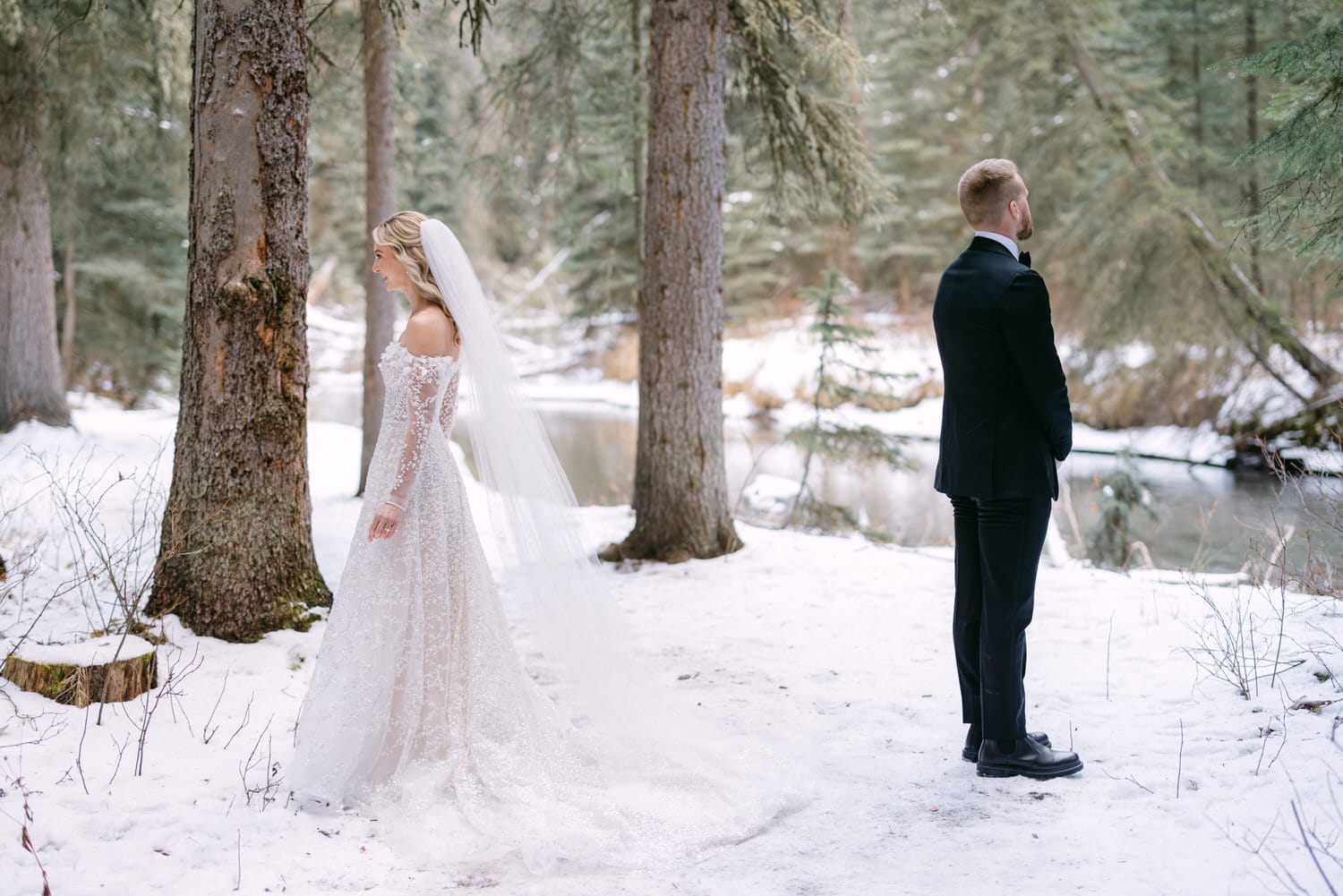 A bride and groom standing back to back in a snowy forest setting, with the bride in a long white dress with a veil and the groom in a classic black suit.