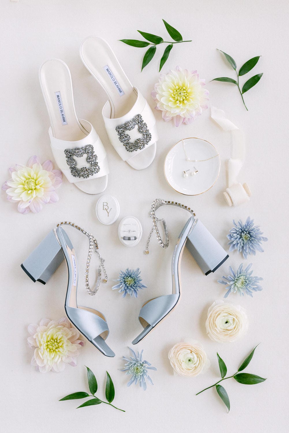 Flat lay of bridal items including white heeled sandals with embellishments, silver jewelry, and fresh flowers on a light background