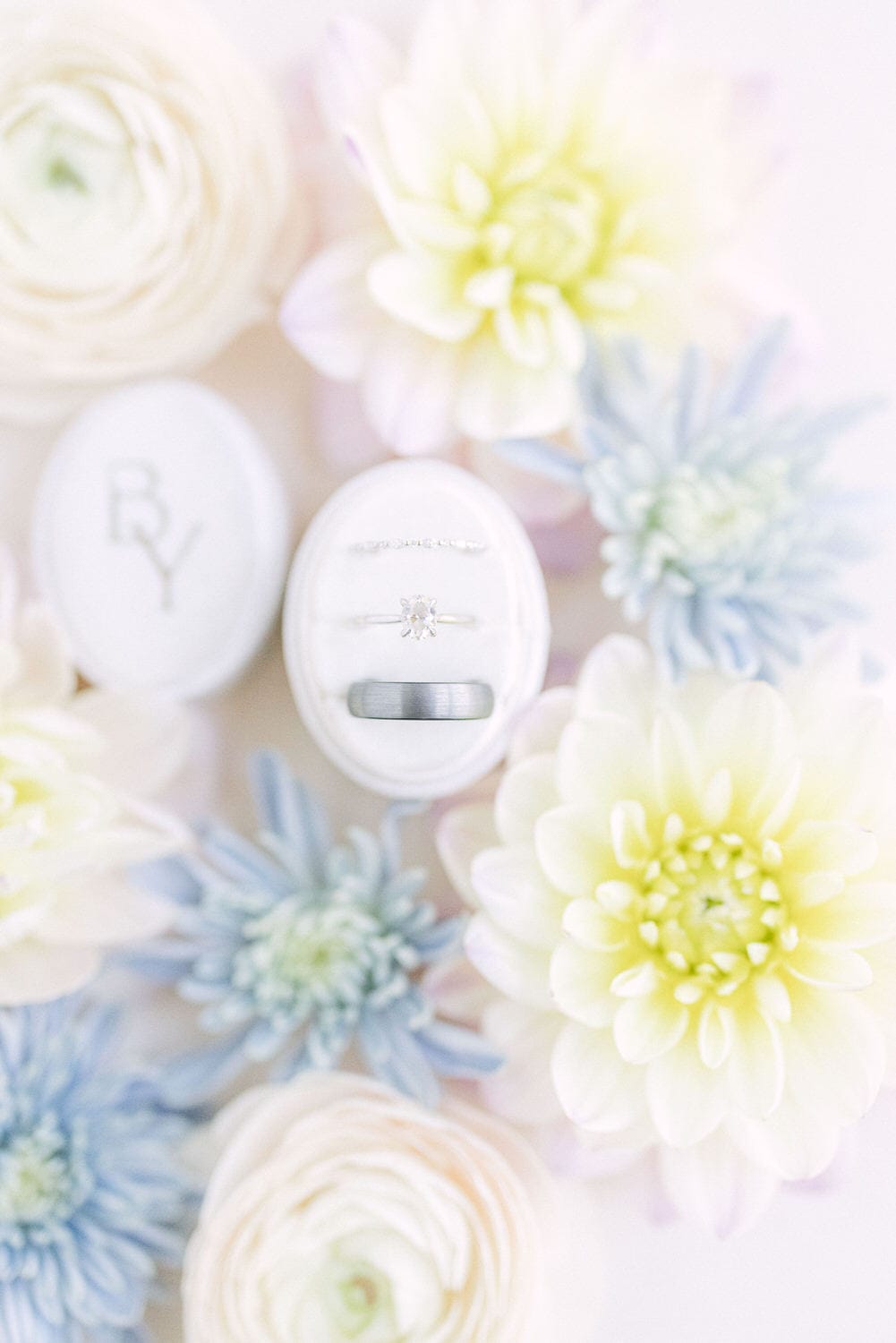 A diamond engagement ring and wedding band displayed in an open ring box surrounded by pastel flowers.