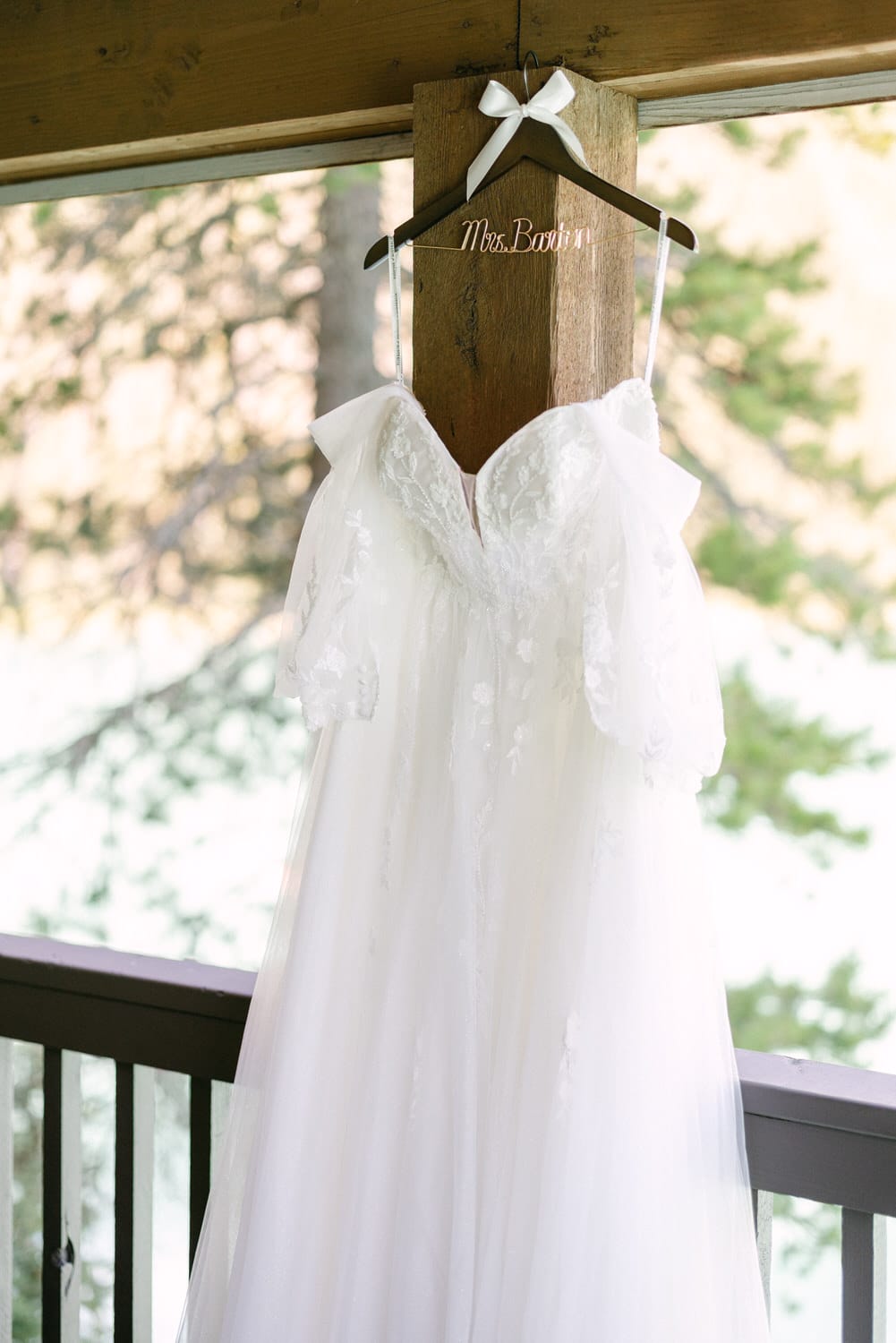 A white wedding dress with lace details hanging from a wooden hanger with "Mrs. Bowen" written on it, displayed on a balcony with a natural backdrop.