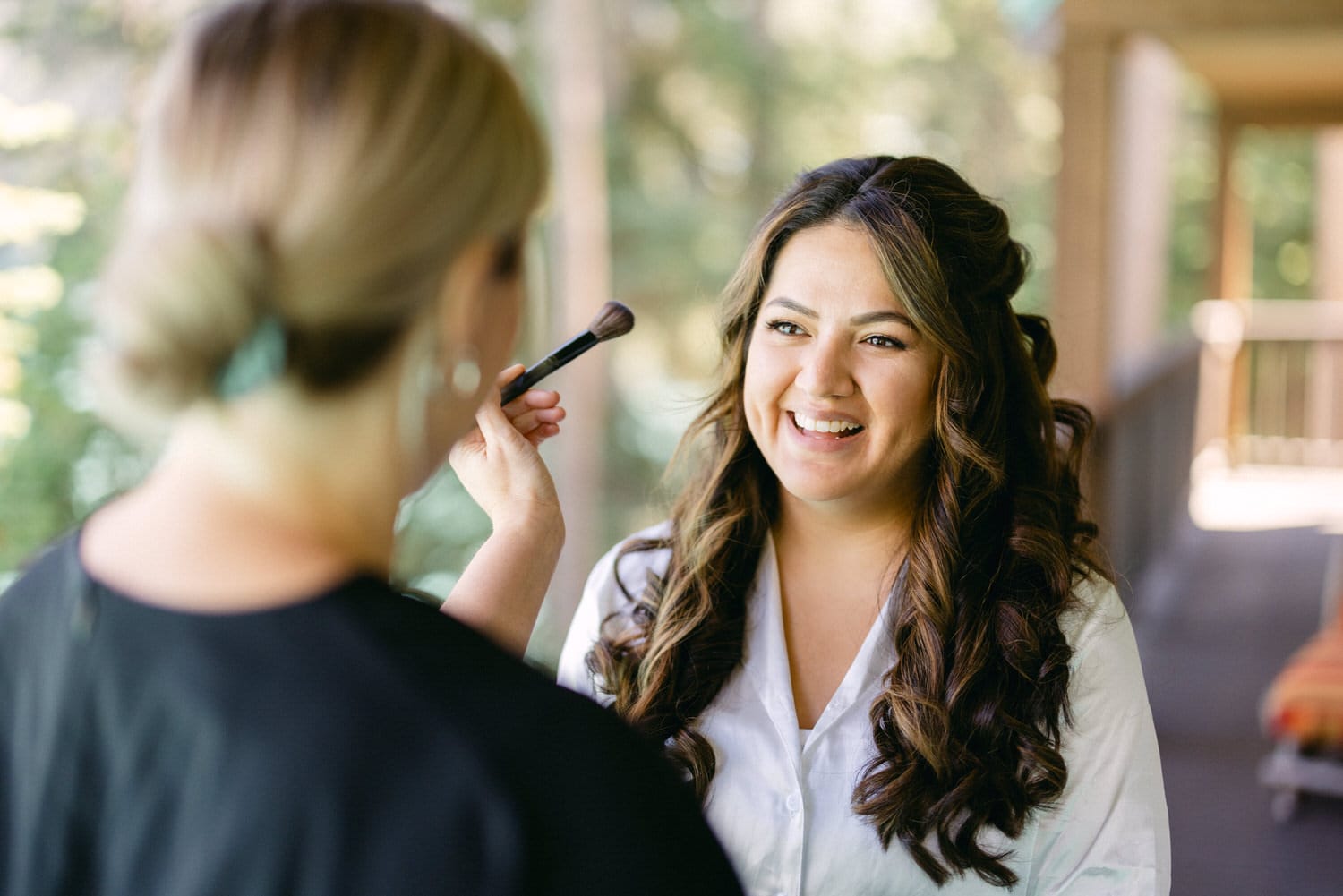 A makeup artist applying makeup to a smiling woman with wavy hair in a natural light setting