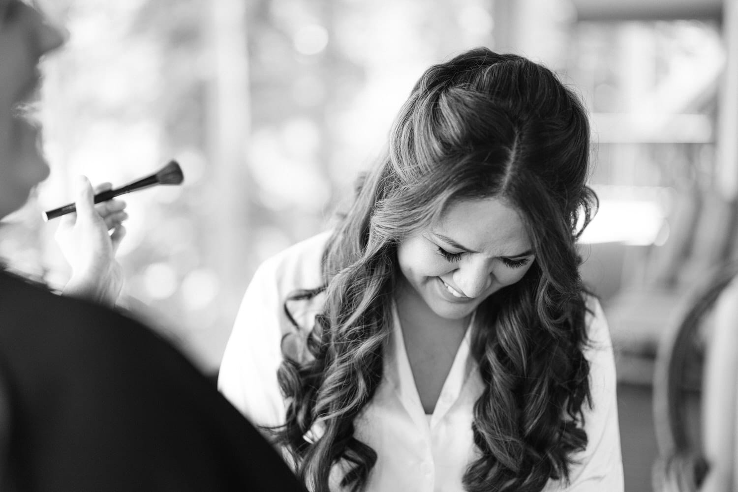 A black and white photo of a smiling bride-to-be getting her makeup done, with a makeup artist applying powder on her face.