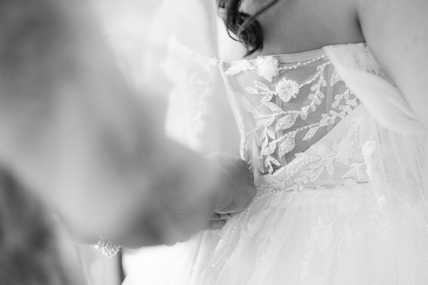 Close-up black and white image of a bride's gown with intricate lace detailing and a hand gently resting on the fabric.