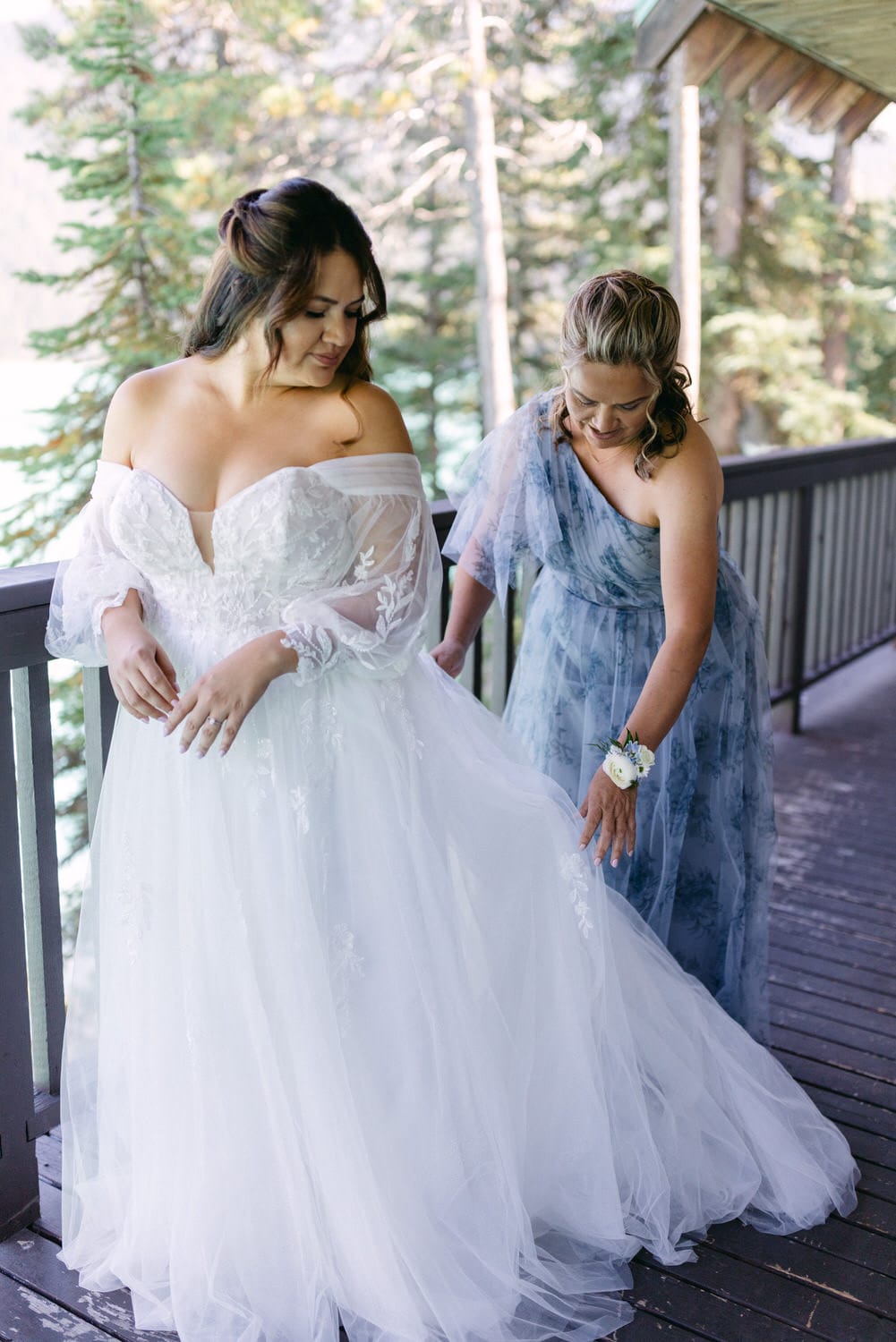 A bride in a white wedding gown being assisted by her bridesmaid wearing a blue dress on a wooden deck, with trees in the background.