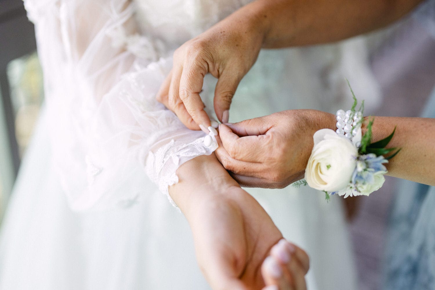 Close-up of hands fastening a white wedding garter on a bride's leg with a floral wrist corsage visible.