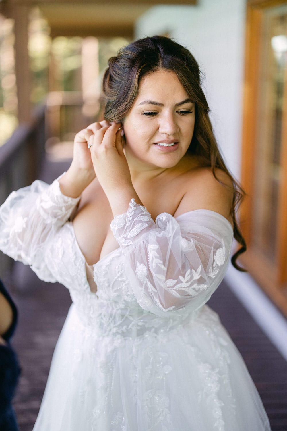 A bride in a beautifully detailed lace wedding dress, touching her earring with a thoughtful expression, against a blurred background.