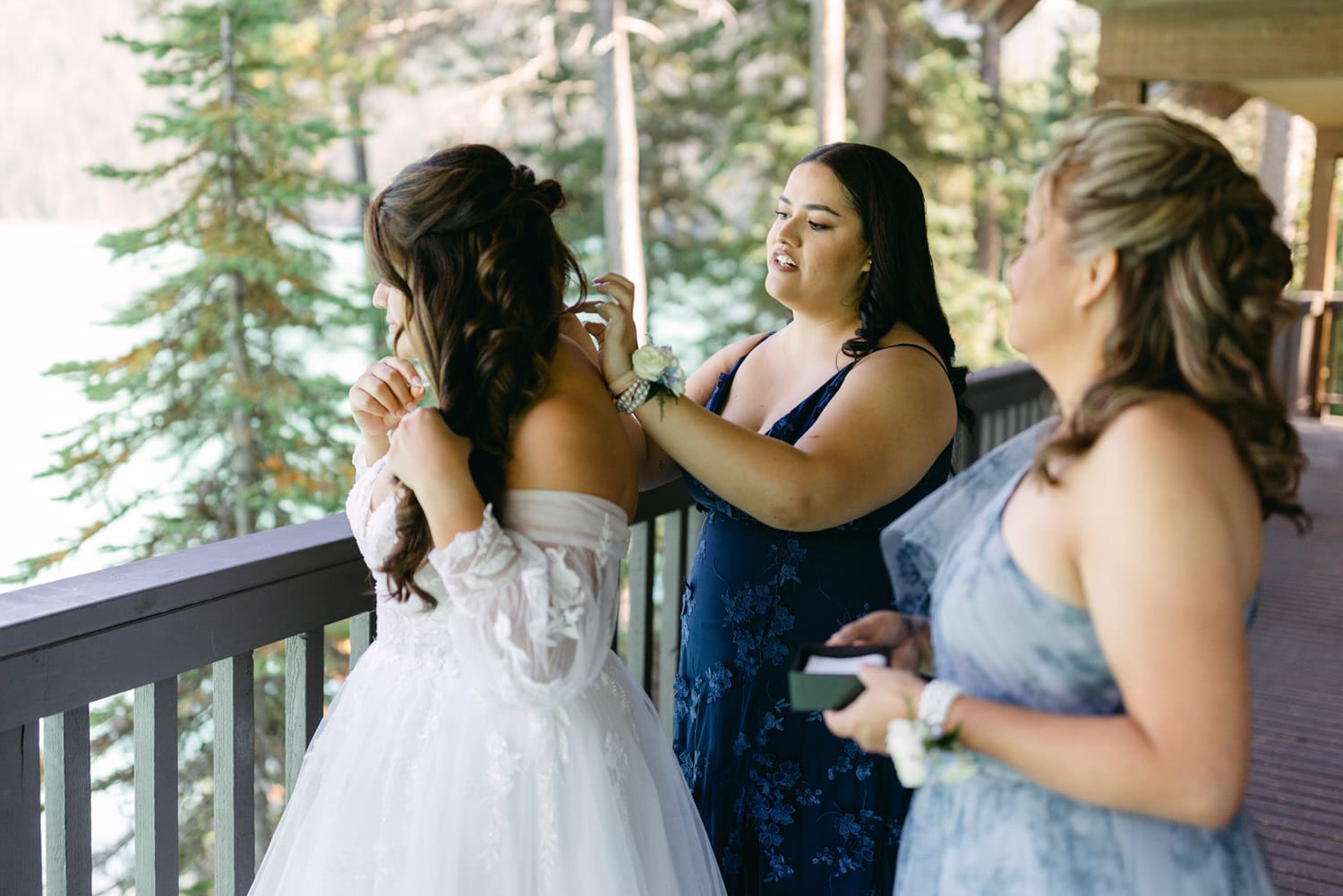 A bride in a white gown being assisted by a bridesmaid in a blue dress while another bridesmaid in grey observes, set against a backdrop of a forest and a lake.