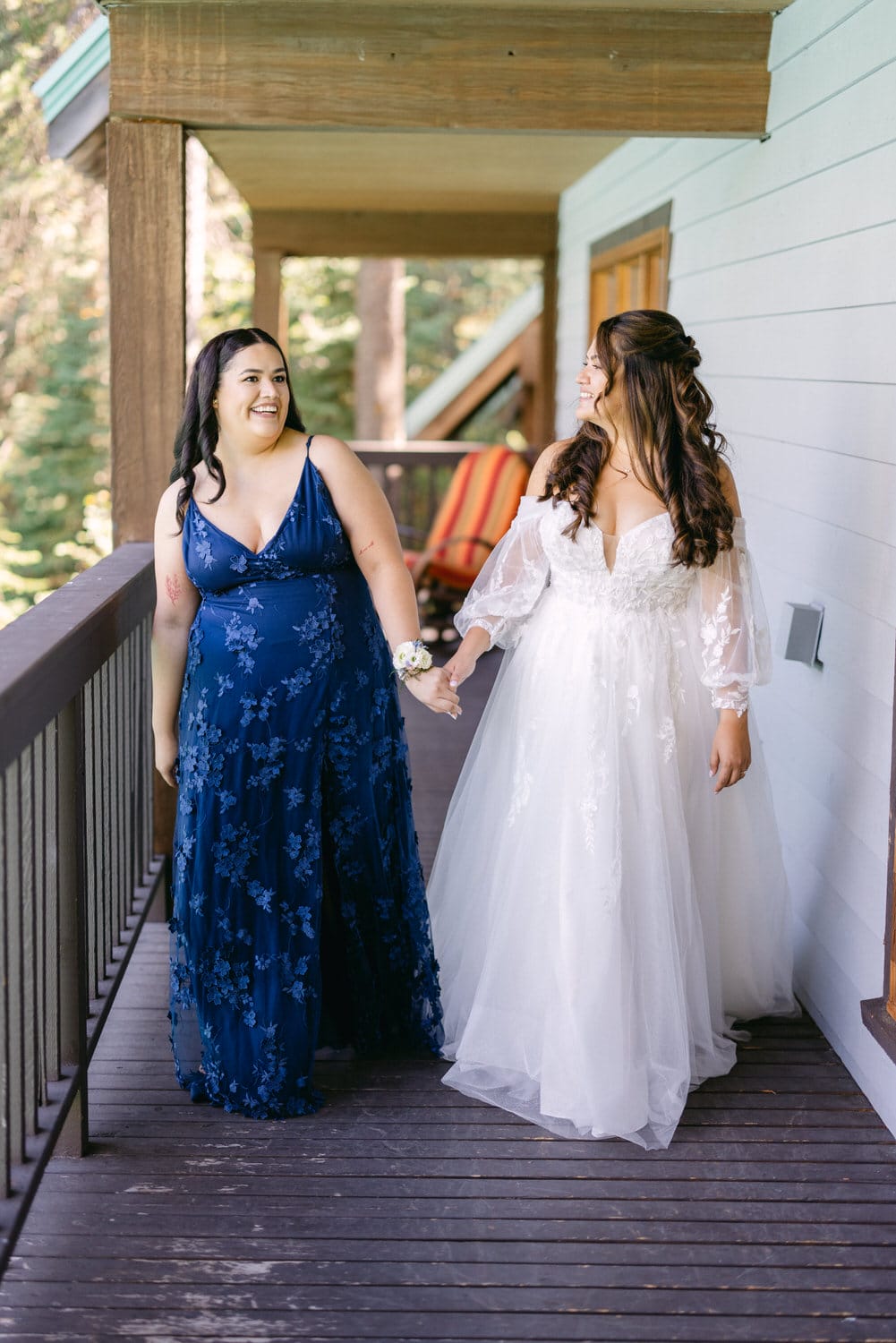 Two women dressed elegantly, one in a blue floral dress and the other in a white bridal gown smiling while holding hands on a wooden deck with a forest background.