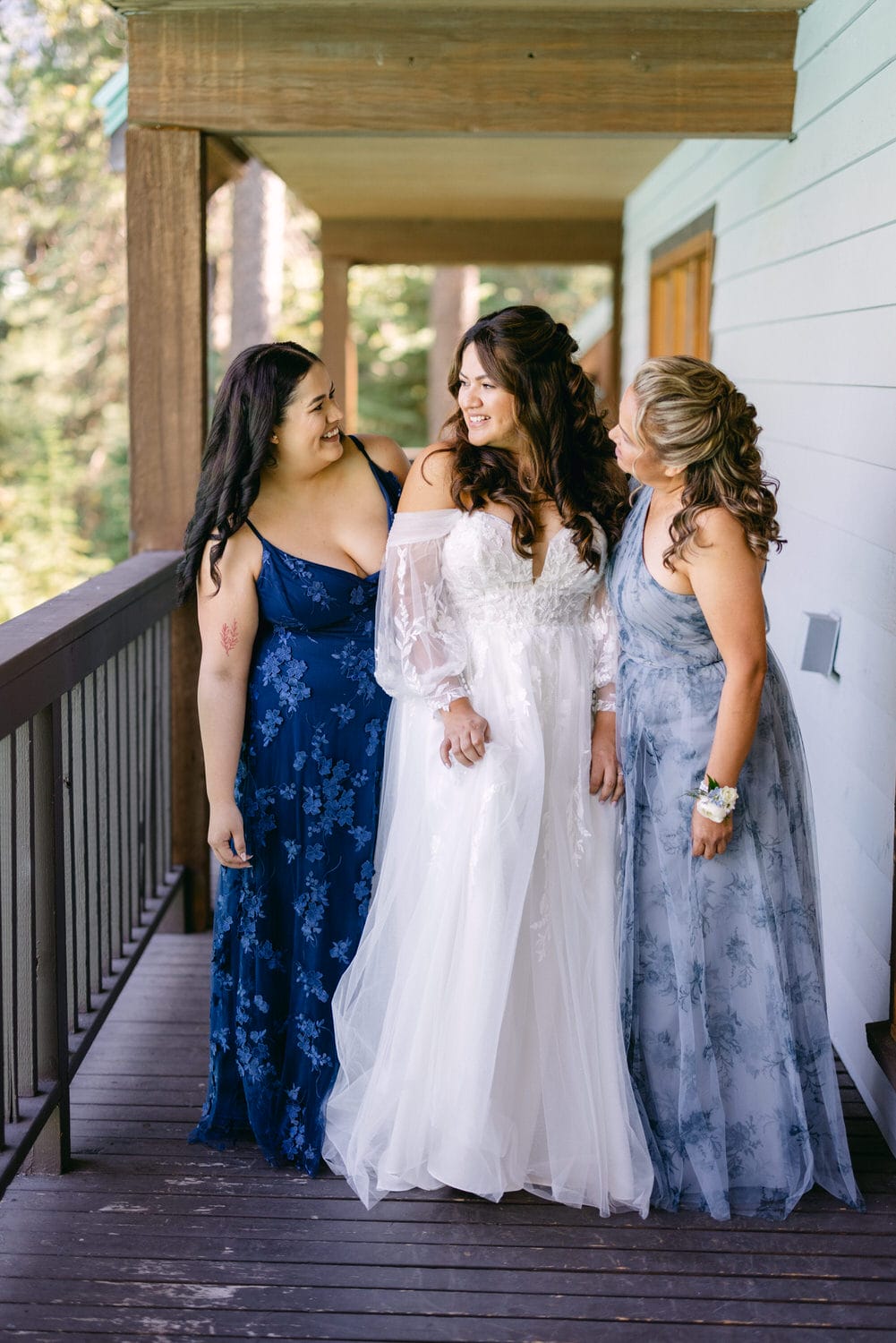 Three women in elegant dresses smiling and sharing a moment together on a wooden deck, with one in a bridal gown and the others in blue themed bridesmaid dresses, surrounded by natural greenery.