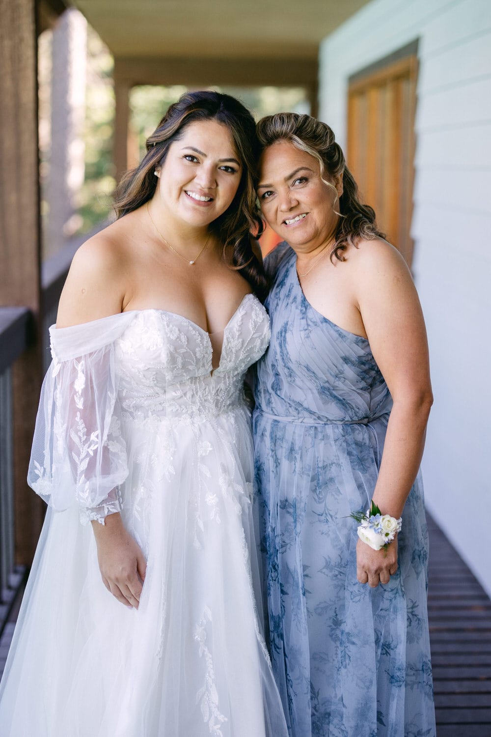 A bride in a white gown smiling alongside her mother wearing a blue dress, both posing for a photo.