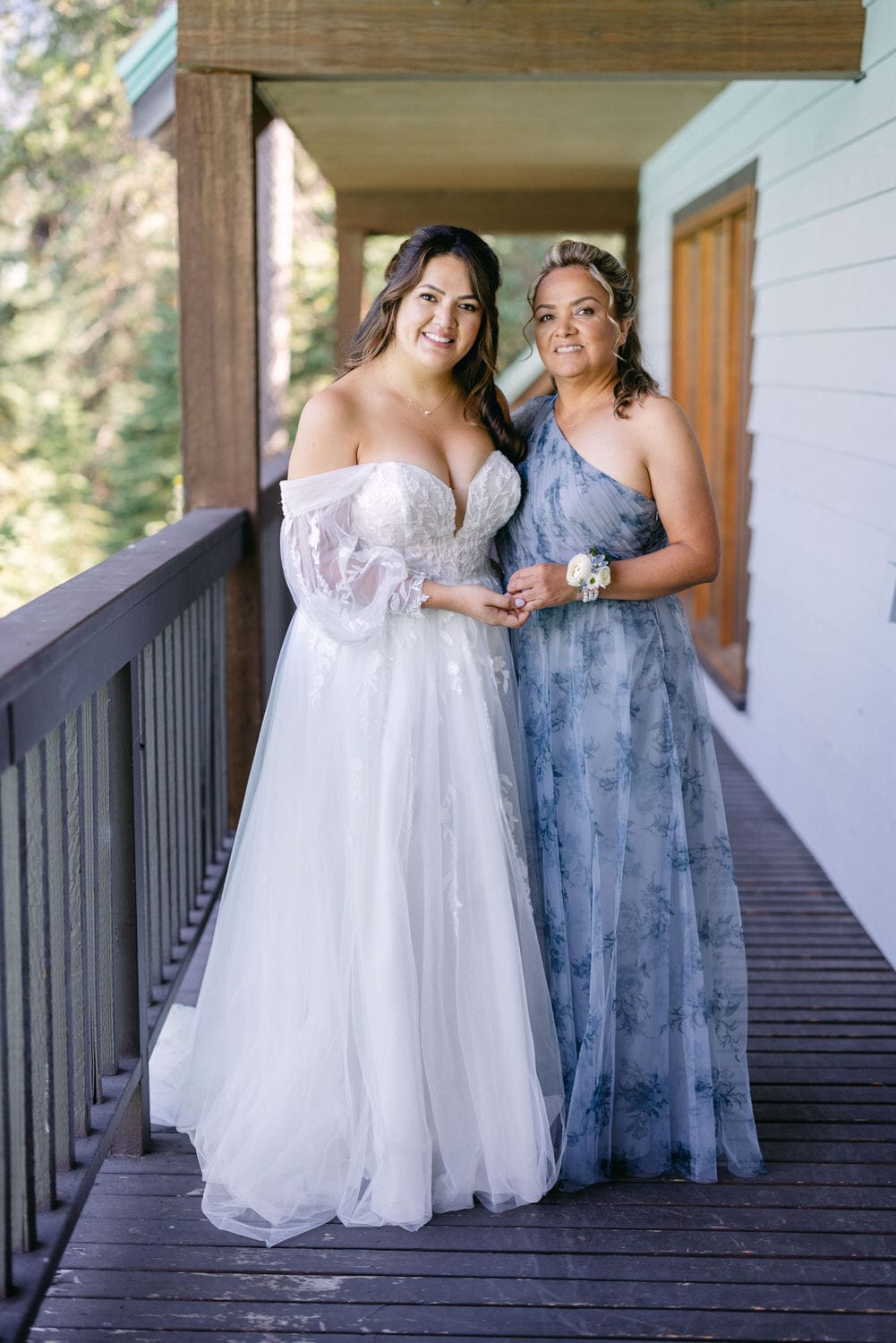 Two women in formal dresses, one in bridal gown and the other in a blue dress, standing together on a wooden deck with a natural backdrop.