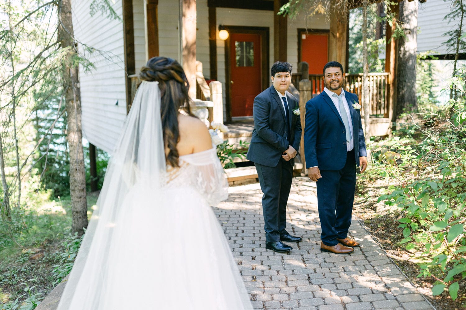 A bride with her back to the camera approaches two men waiting on a cobblestone path, with the groom and best man smiling towards her.