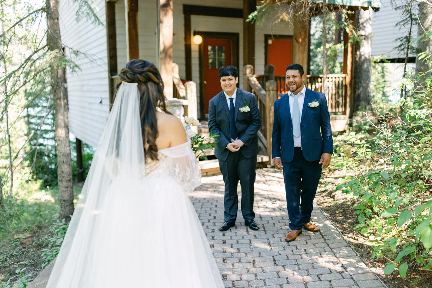 A bride with her back towards the camera looking at the smiling groom and best man approaching on a wooded path.