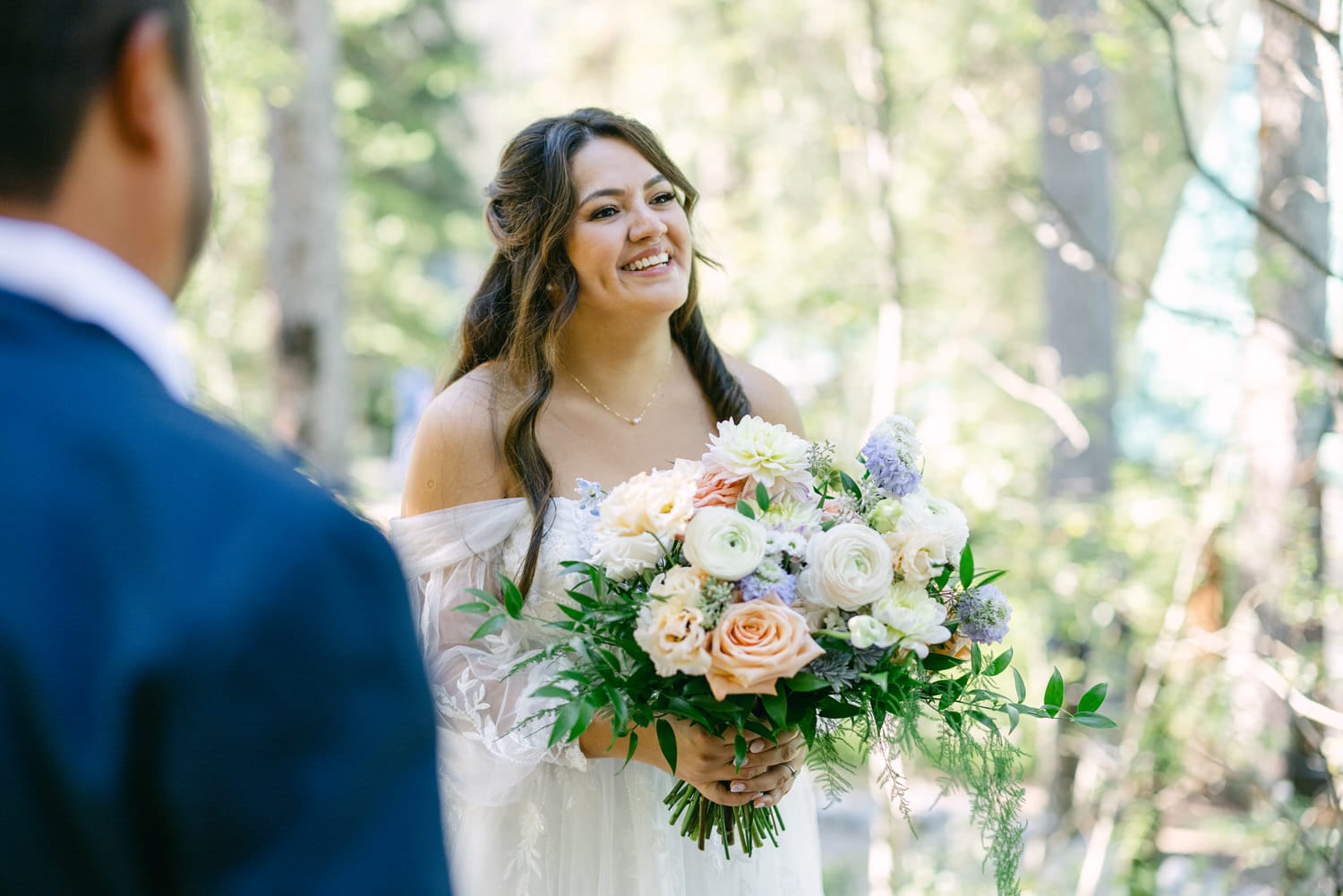 A smiling bride in an off-shoulder dress holding a delicate bouquet of flowers with a blurred groom in the foreground and a serene forest backdrop.