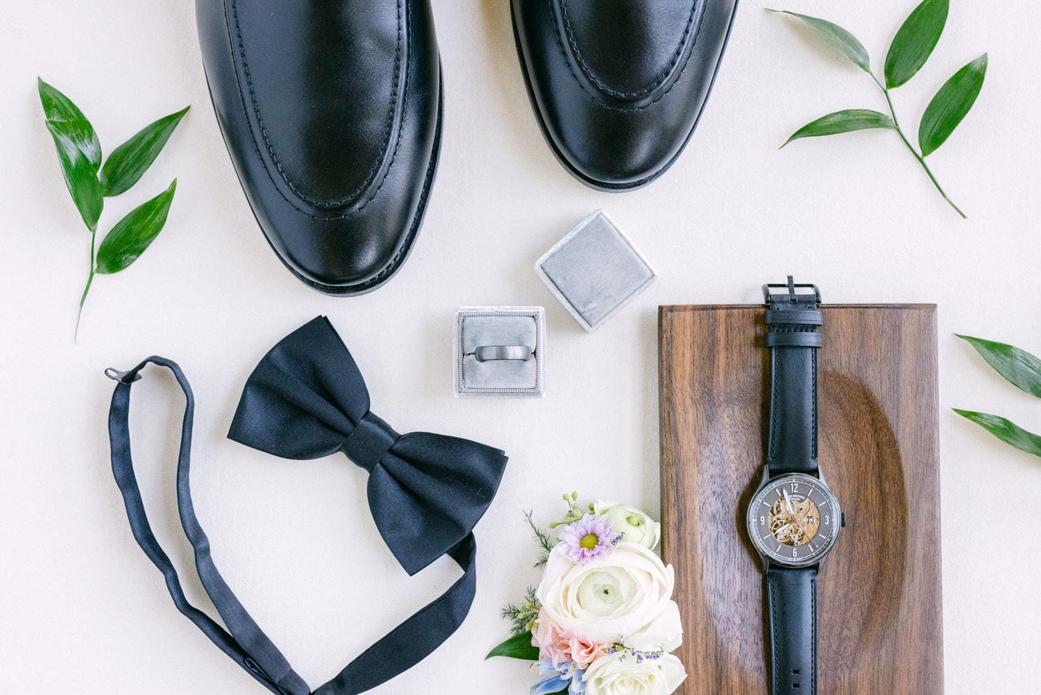 Top view of men's wedding accessories including black shoes, bow tie, cufflinks, wristwatch, and a small floral bouquet on a neutral background.