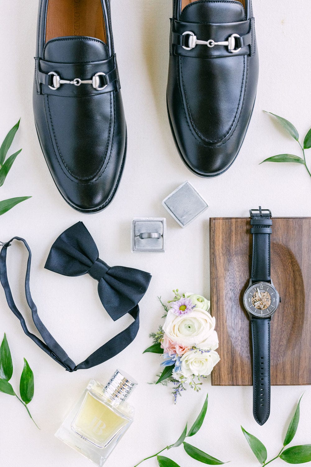 A pair of black dress shoes, bow tie, cufflinks, wristwatch, boutonniere, and cologne bottle arranged neatly on a light background with green leaves.