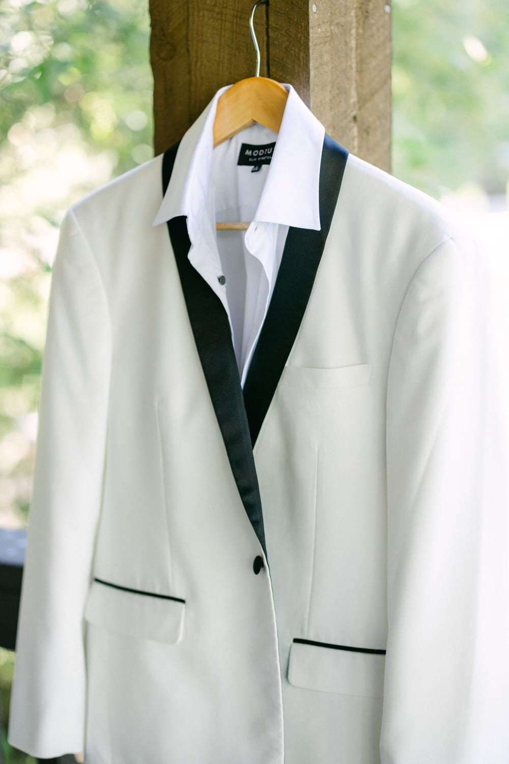 A white tuxedo with black lapels and a white dress shirt hanging on a wooden hanger outdoors