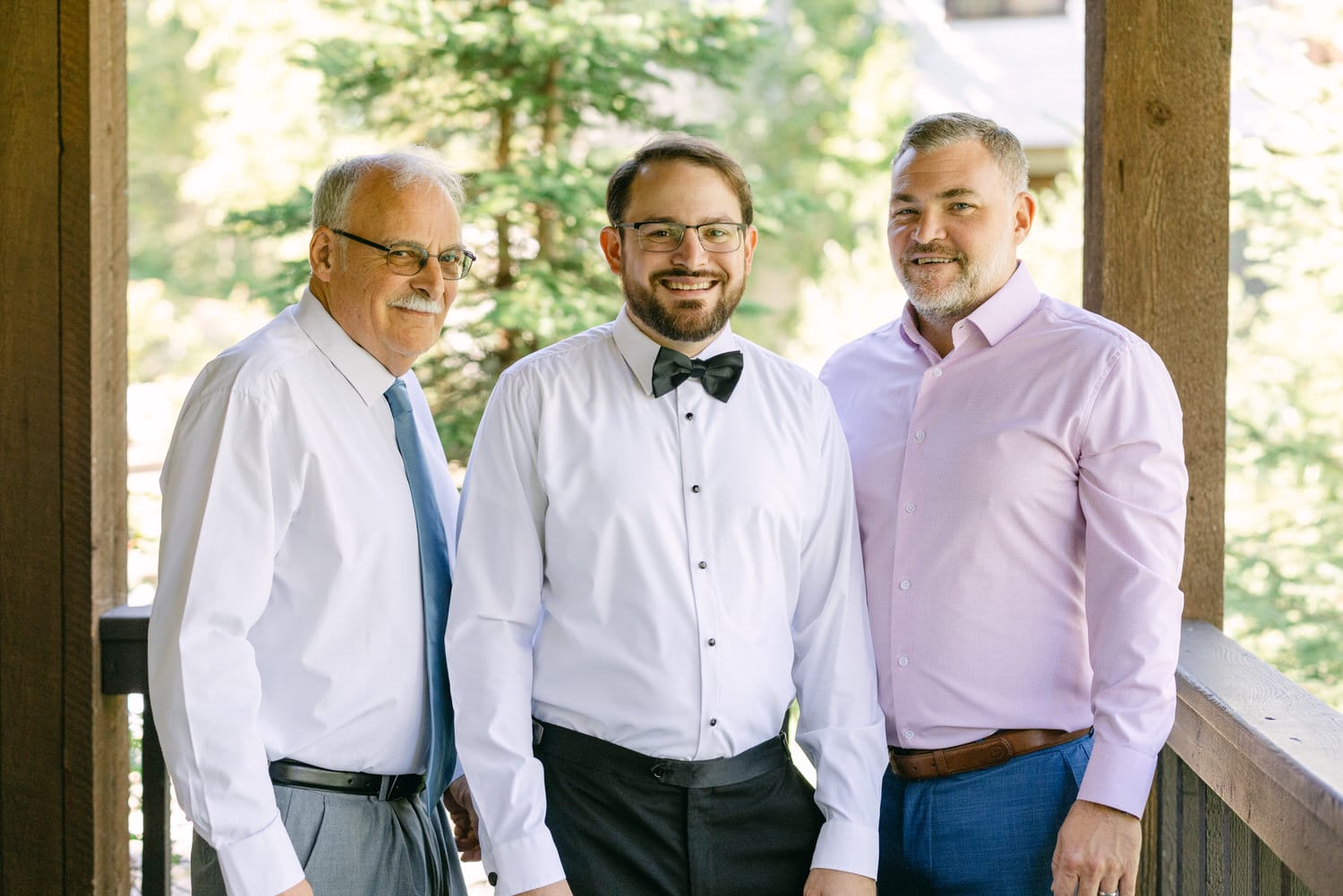 Three smiling men of different ages standing together on a wooden porch, surrounded by trees.