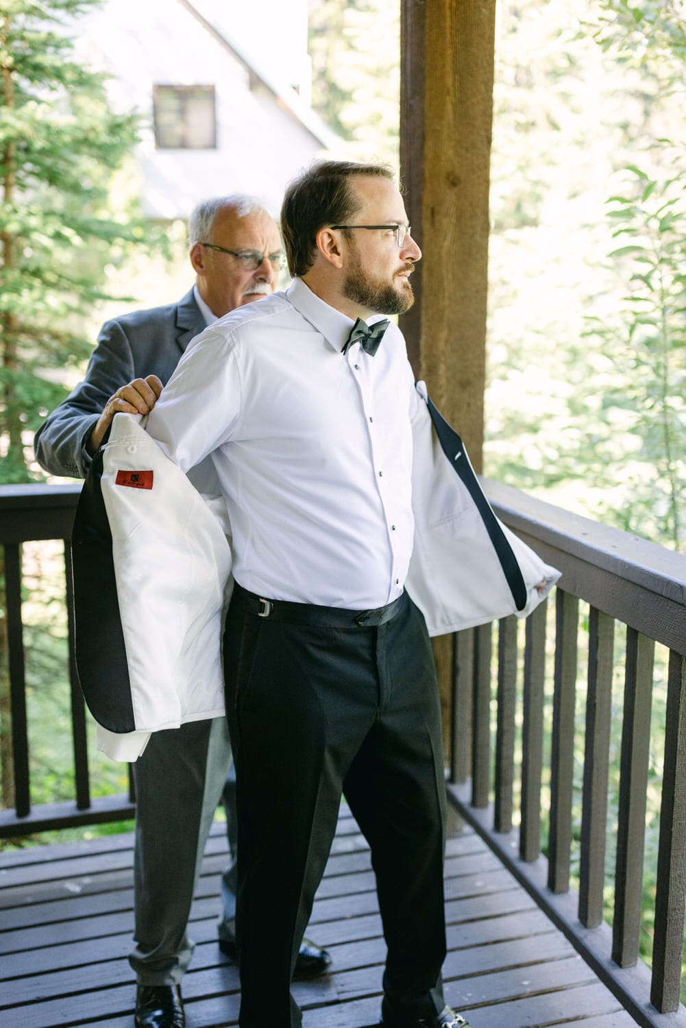 An older man assists a younger man with his suit jacket on a wooden balcony, both dressed in formal attire for a special occasion.