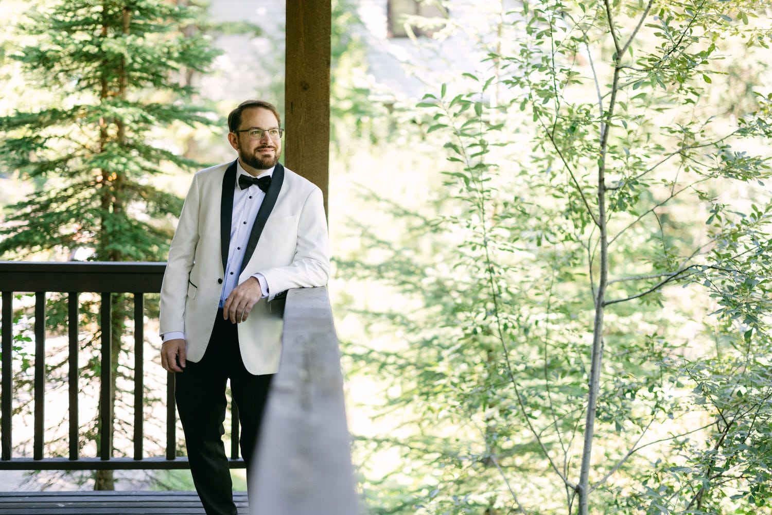 A man in a white tuxedo jacket and black bowtie leaning on a wooden railing amidst lush greenery.