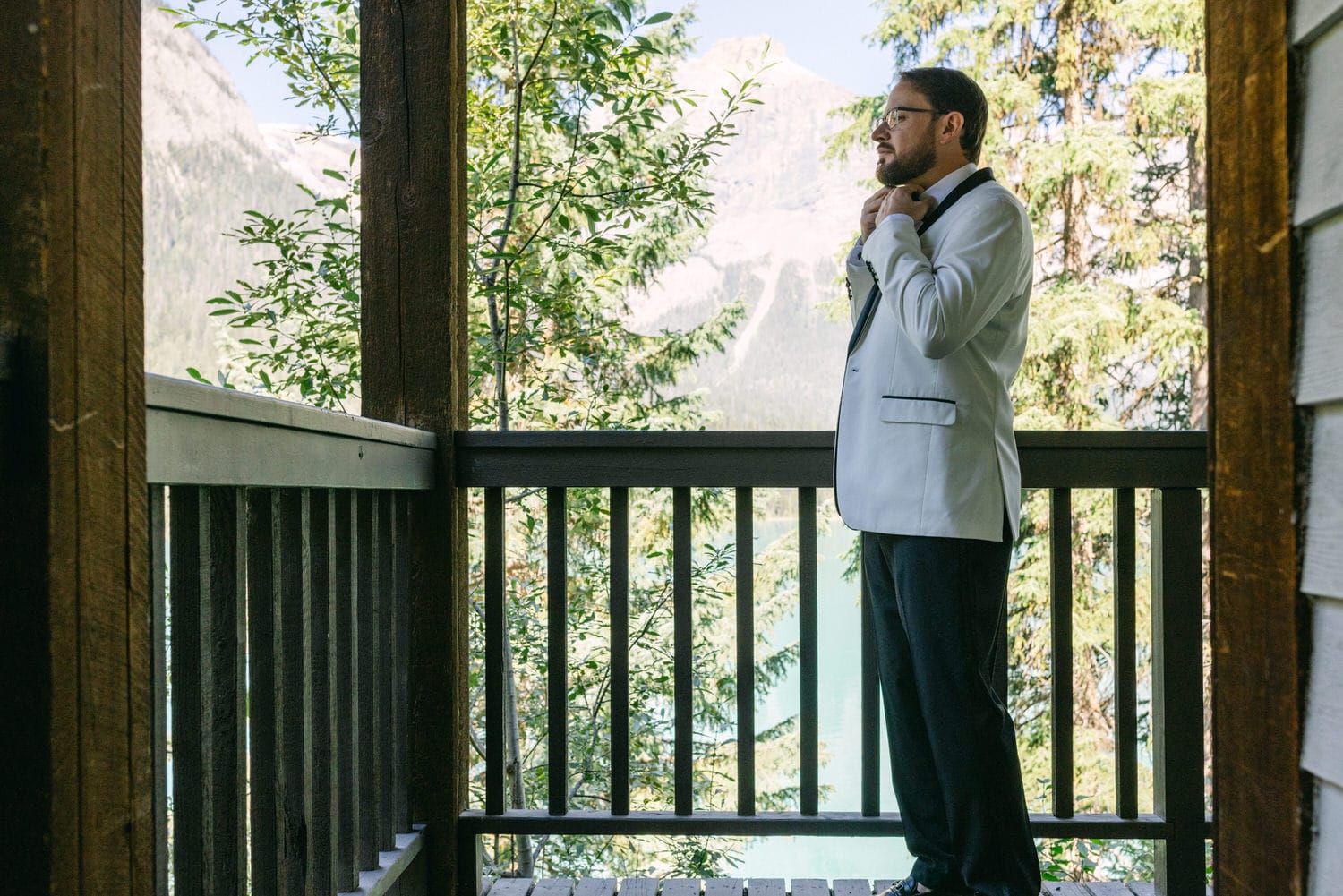 A man in a formal white jacket and black trousers is adjusting his tie while standing on a balcony with lush greenery and mountain views in the background.