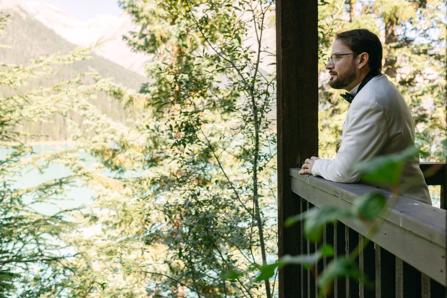 A man in a white shirt and glasses leaning on a wooden railing, looking thoughtfully at a scenic forest and mountain landscape.