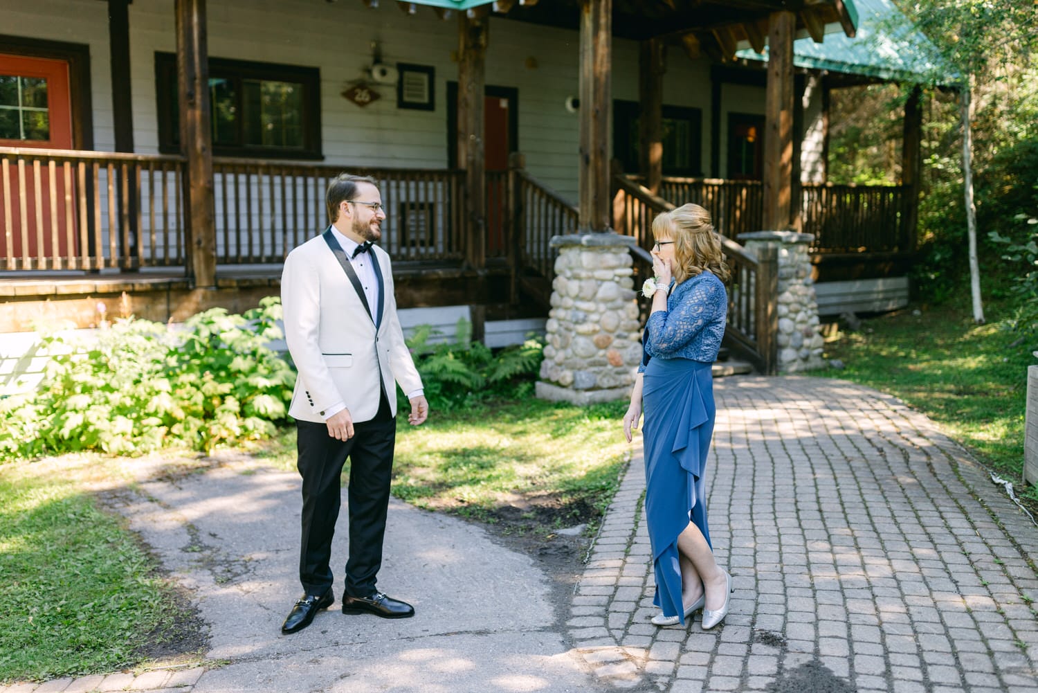 A man in a white tuxedo and a woman in a blue dress standing on a cobblestone path outside a rustic wooden cabin with greenery around.