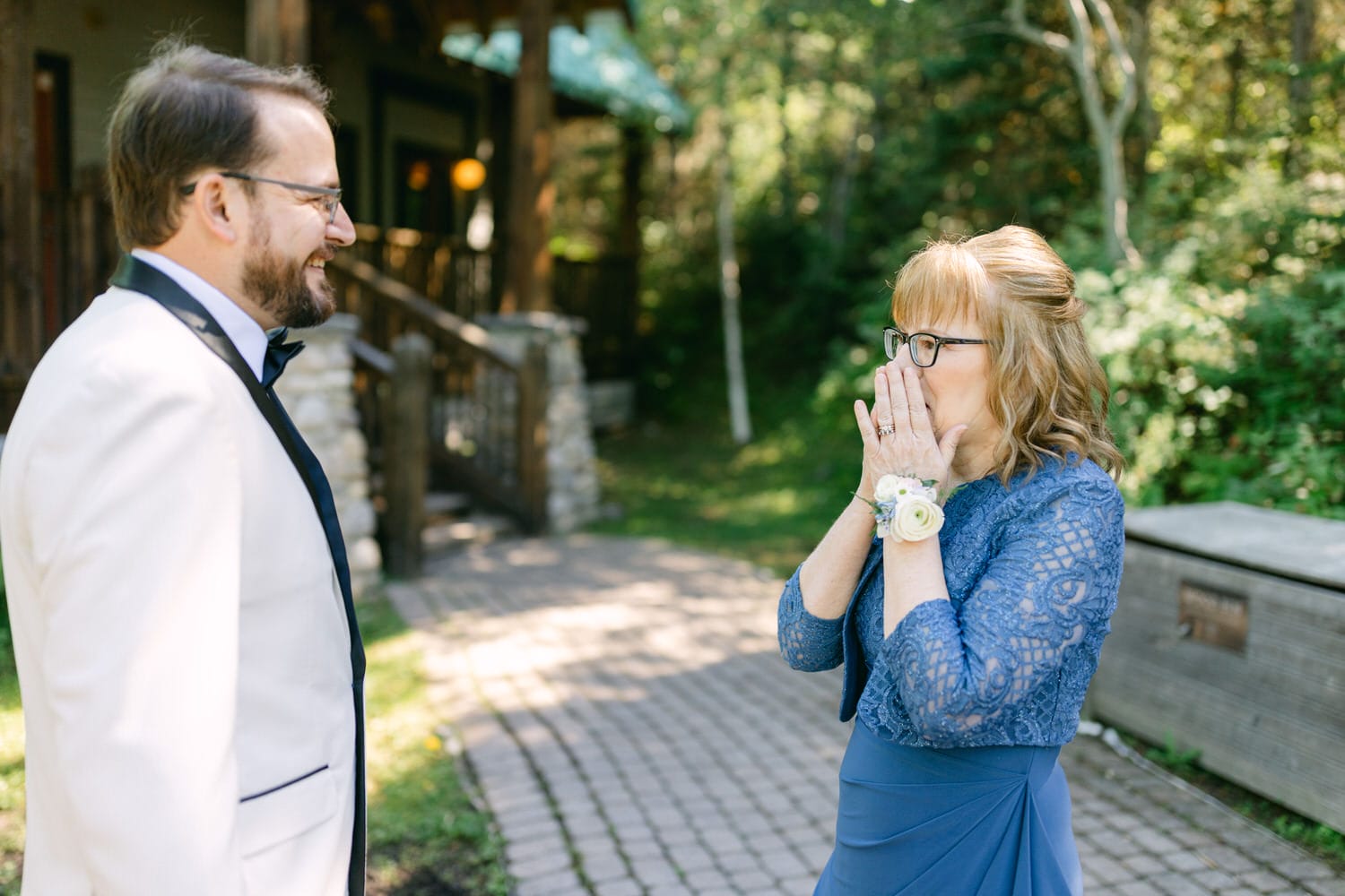 A woman in a blue dress expressing surprise while covering her mouth with hands as she looks at a man in a white suit smiling at her. They are standing outdoors near a building with greenery in the background.