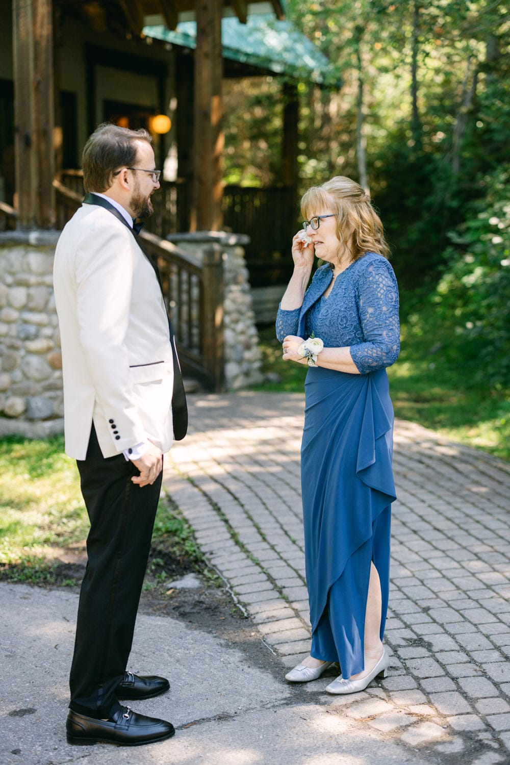 A woman wiping away a tear while talking to a man in a white jacket in front of a rustic building surrounded by trees.