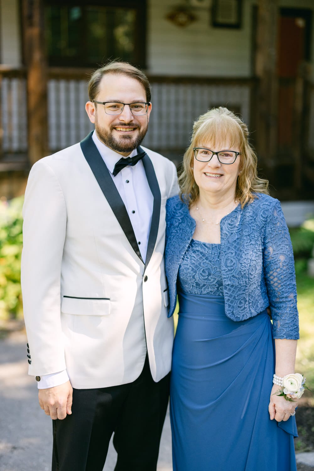 A man in a white tuxedo and a woman in a blue dress with a lace jacket, both smiling, in front of a rustic wooden building.