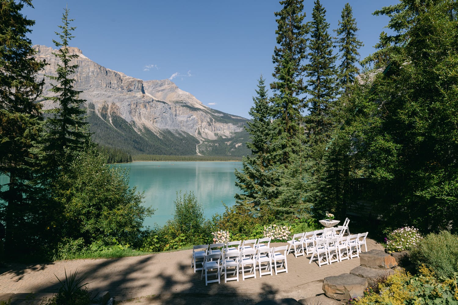 Rows of white chairs arranged for a wedding ceremony overlooking a serene turquoise lake with majestic mountains and lush trees in the background.