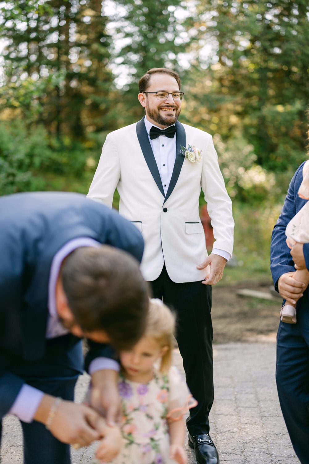 A beaming groom in a white jacket and black bow tie at an outdoor wedding, with a young girl in a floral dress in the foreground and another guest partially visible.