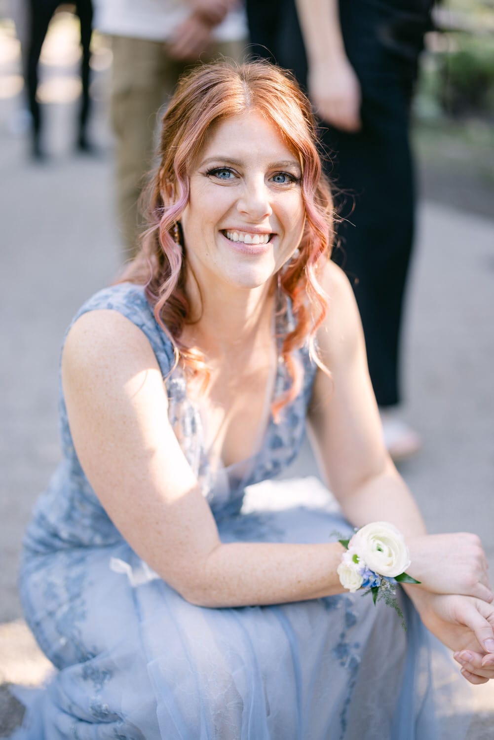 A smiling woman with pink hair wearing a blue dress and a wrist corsage outdoors.