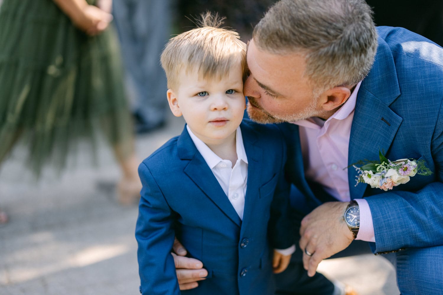 A man in a blue suit kissing a young boy in a matching blue suit on the cheek, both at an outdoor event.