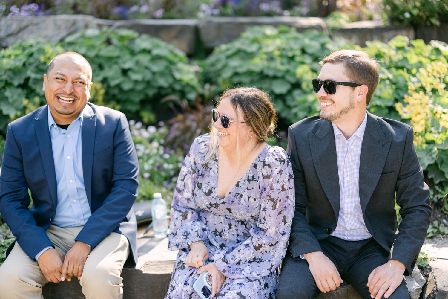 Three people smiling and sitting outdoors in a garden with greenery and flowers in the background. The person on the left and the person in the middle are wearing sunglasses.