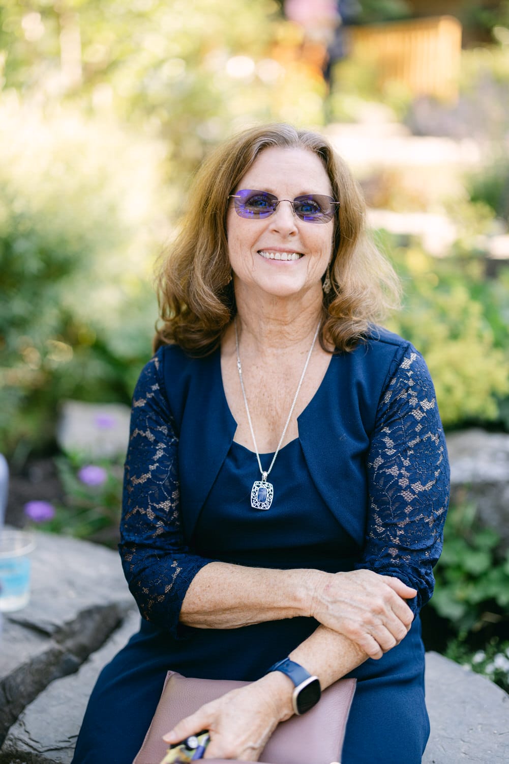 A smiling senior woman with sunglasses and a blue dress seated on a rock in a garden setting.