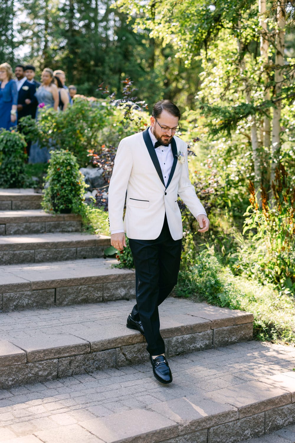 A man in a white tuxedo jacket and black trousers walking down stone steps at an outdoor wedding with guests in the background