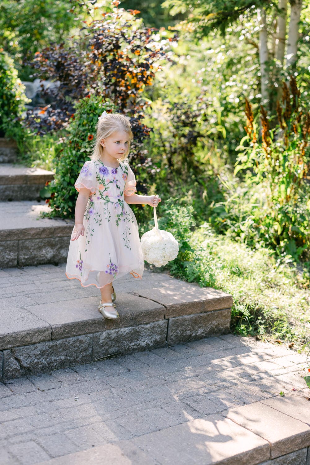 A young girl in a floral dress holding a flower ball while walking down garden steps.