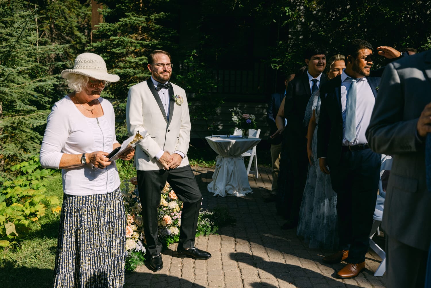 A woman in a white hat reading to a smiling groom at an outdoor wedding with guests looking on.