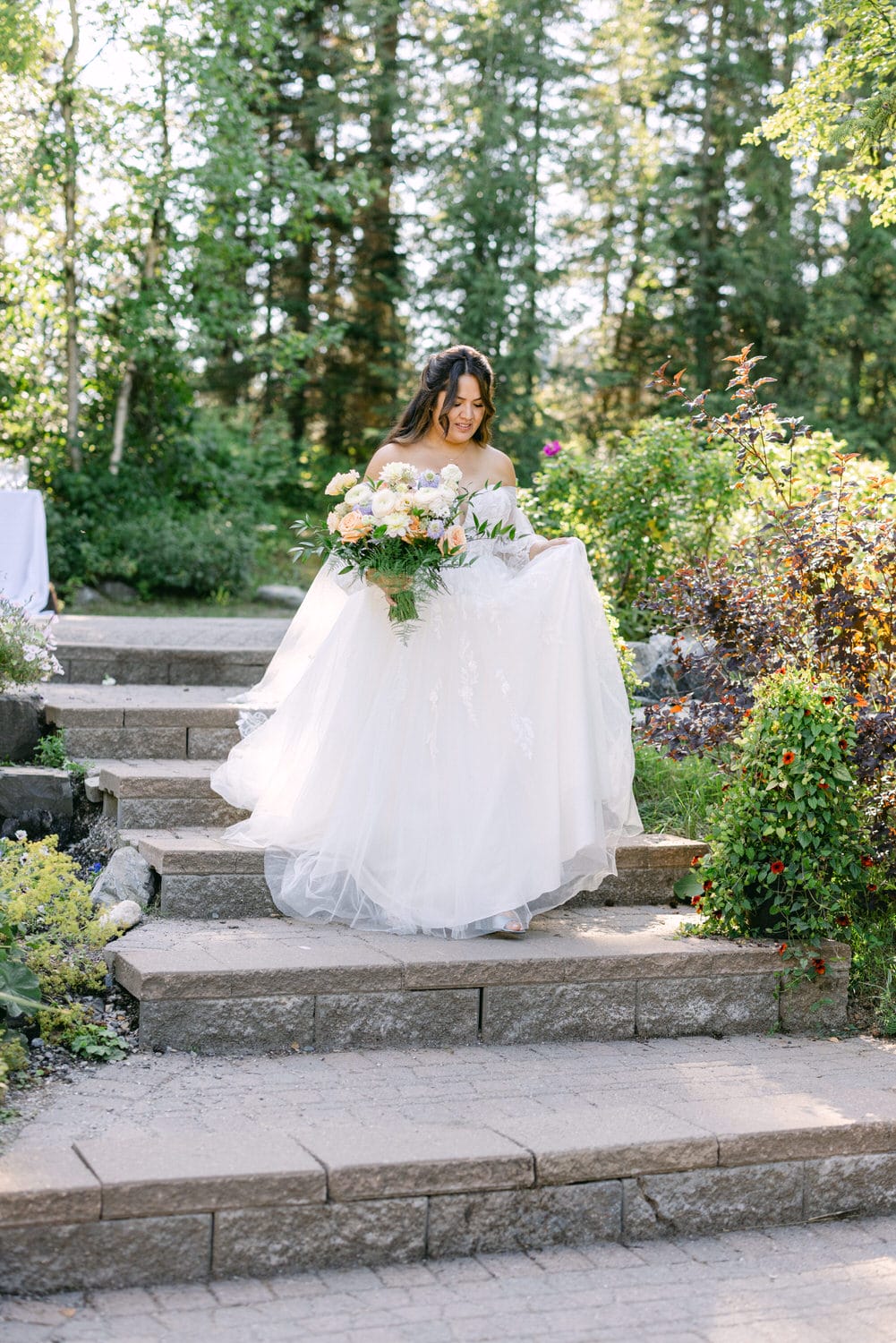 A bride in a white wedding dress carrying a bouquet while walking down an outdoor staircase surrounded by lush greenery.