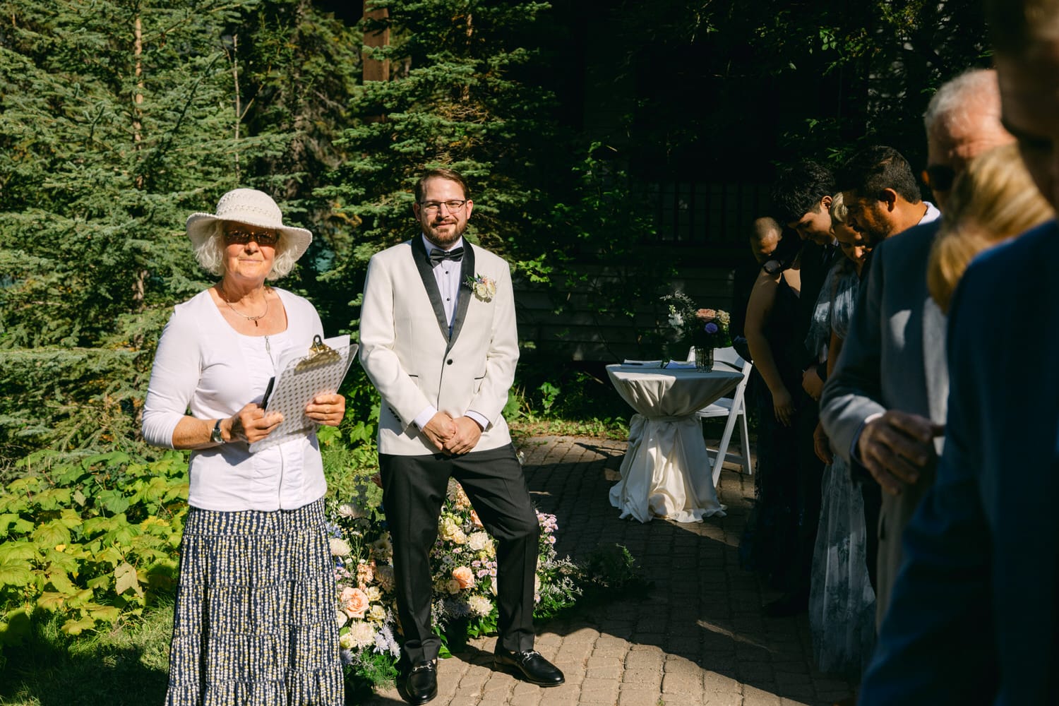 A groom standing with anticipation alongside wedding guests at an outdoor ceremony setting, with a woman in a white hat holding a program in the foreground.