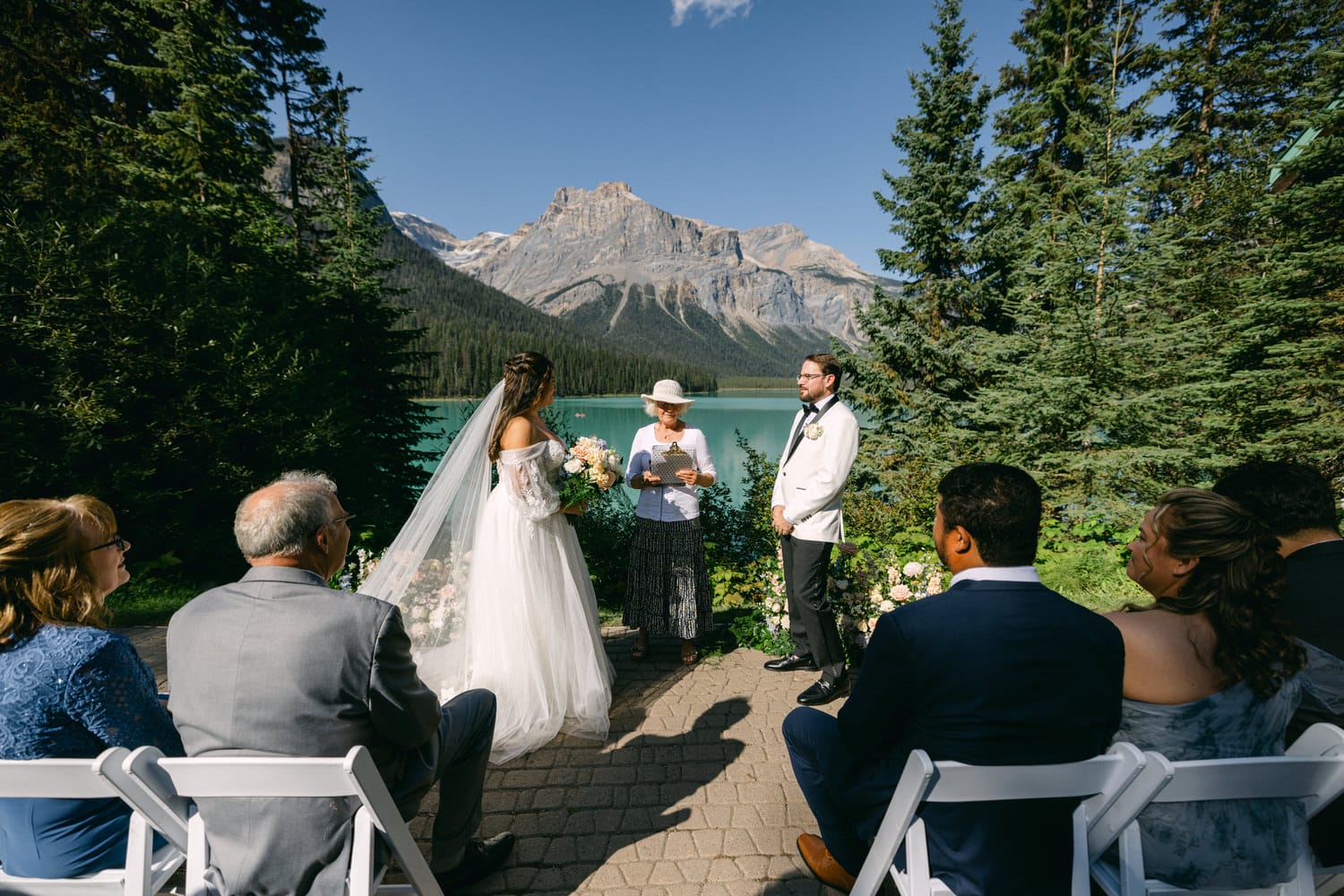 A bride and groom stand at the altar with a scenic mountain lake in the background, surrounded by guests at their outdoor wedding ceremony.