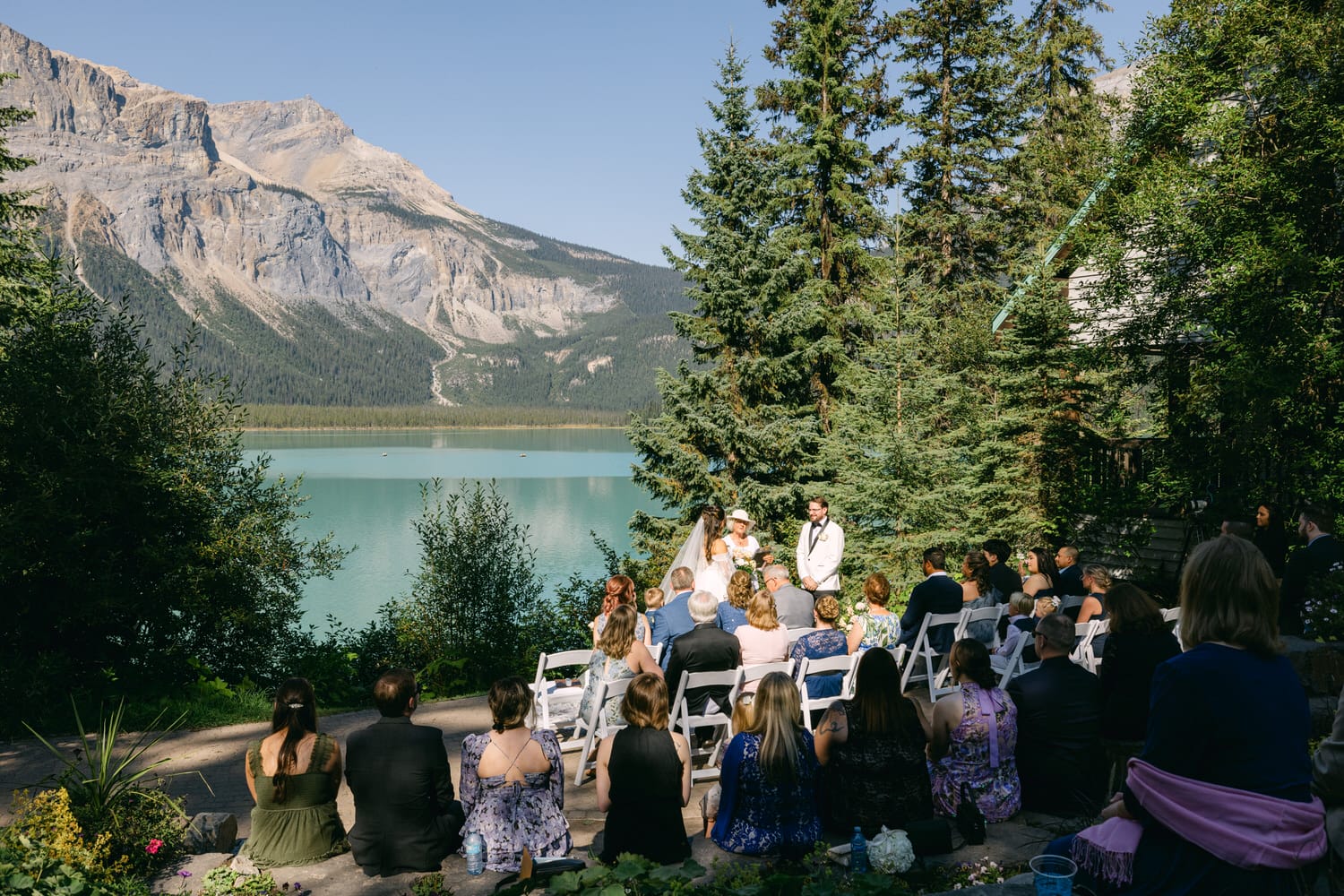 A couple exchanging vows at an outdoor wedding ceremony with guests seated, surrounded by lush greenery and a scenic mountain lake backdrop.