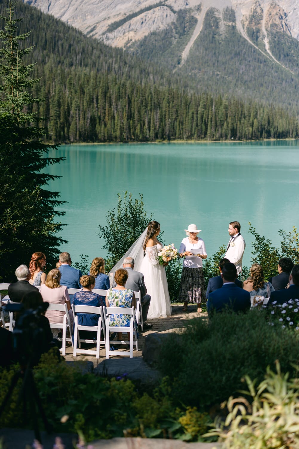 A bride and groom stand before an officiant at an outdoor wedding ceremony by a turquoise lake, with guests seated and mountains in the background.