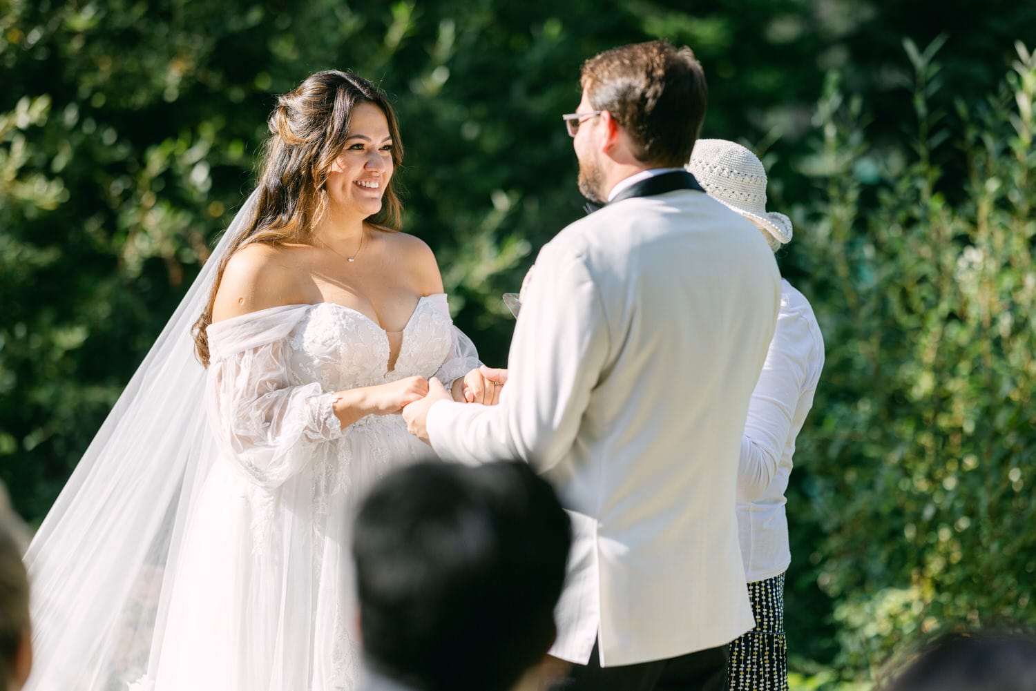 A bride and groom holding hands and smiling at each other during a wedding ceremony outdoors, with greenery in the background.