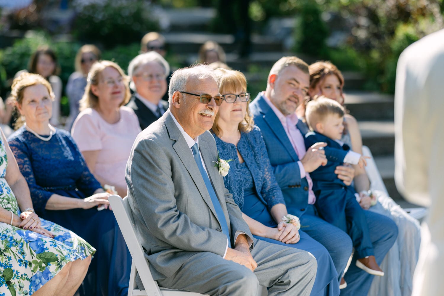 A group of guests seated at a wedding ceremony outdoors with focused attention towards the front.