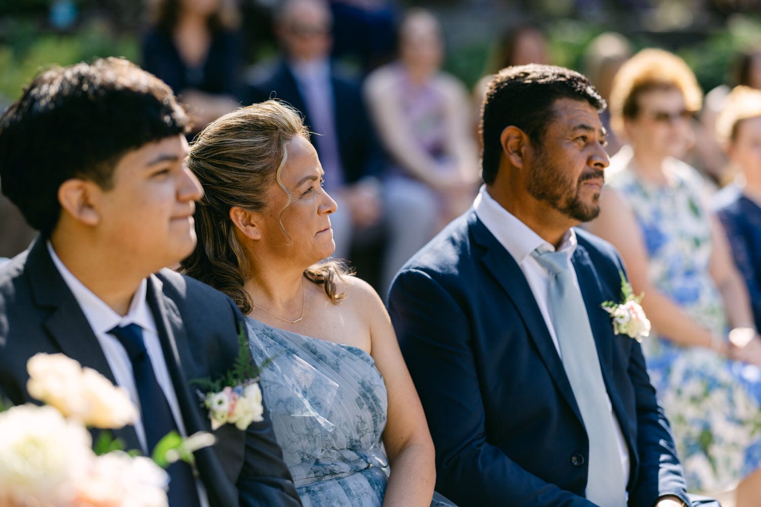 Three guests in formal attire seated at an outdoor event, attentively watching the proceedings with a crowd in the background.