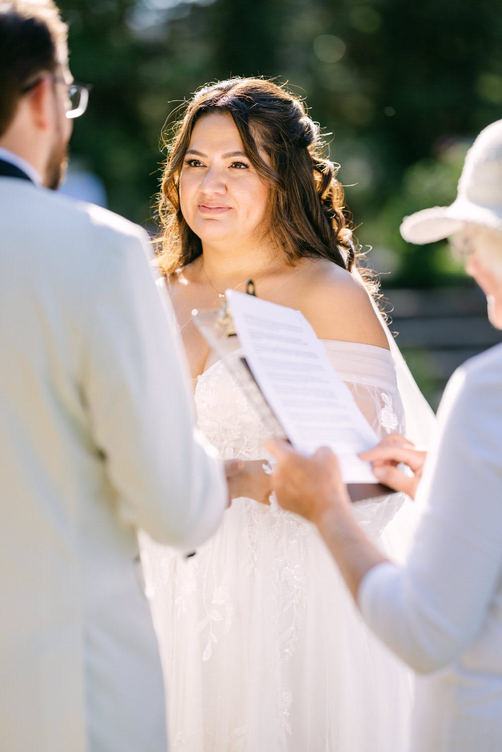 A bride in an off-shoulder wedding dress exchanging vows with a groom, as an officiant holds the vows on a clipboard, in a sunlit outdoor setting.