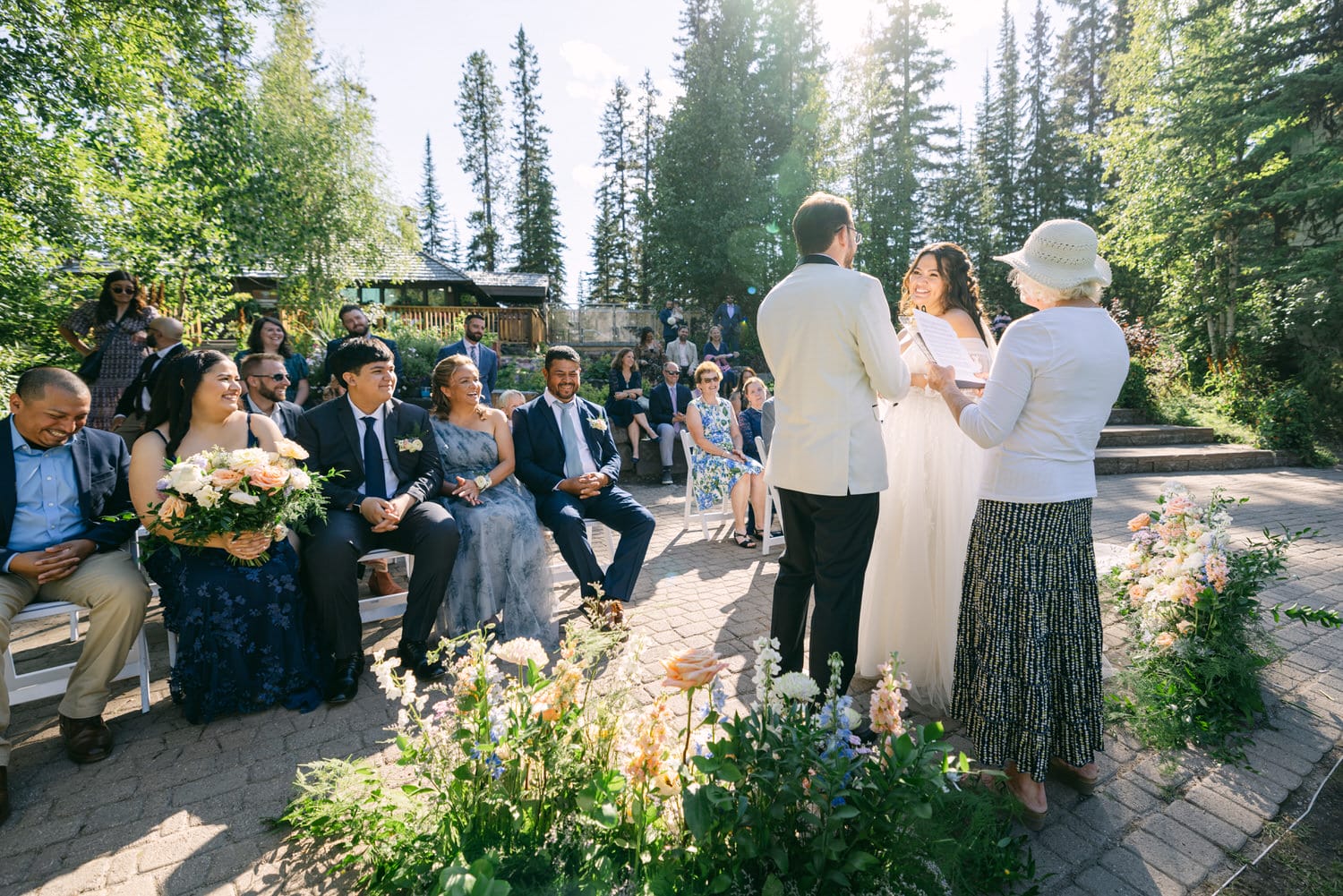 Bride and groom exchanging vows at an outdoor wedding with guests seated on either side and flowers lining the aisle.