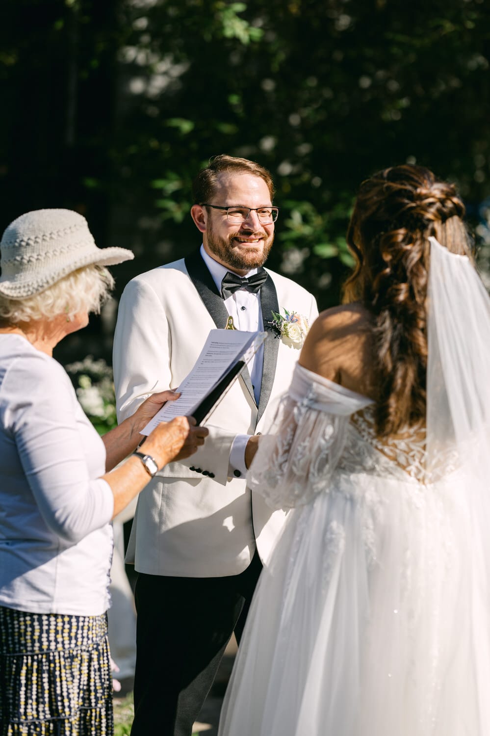 Groom smiling while looking at bride, with officiant holding a book in the foreground during an outdoor wedding ceremony.